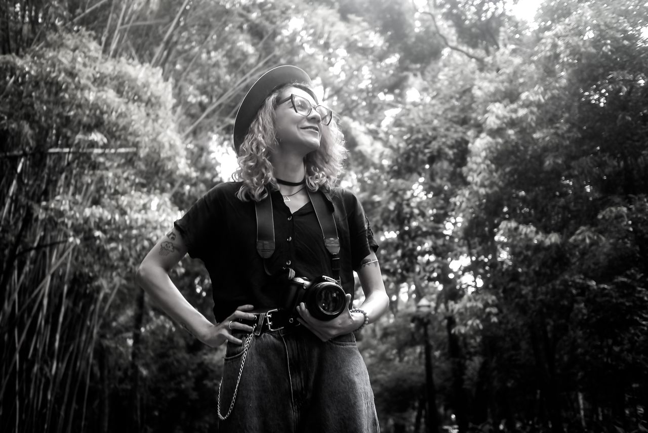 Services Black and white portrait of a confident photographer with her camera in a lush outdoor setting.